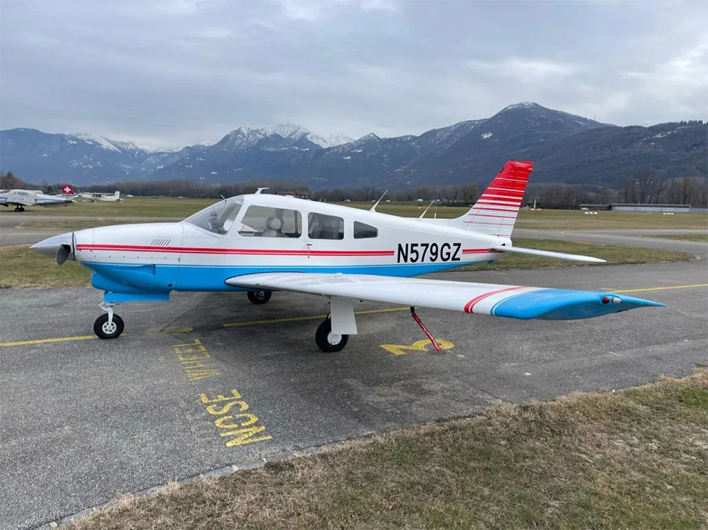 A white and blue Piper PA-28-181 Archer II with red stripes on the tail and wings, parked on an airfield with mountains in the background.