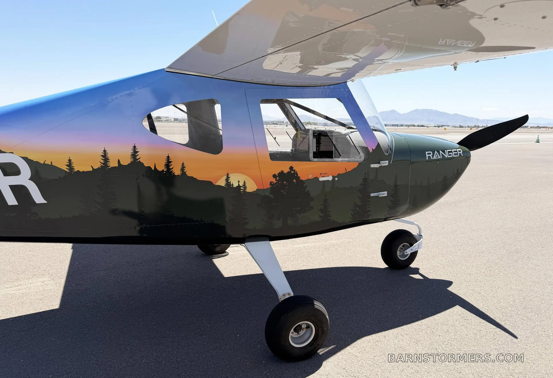 A close-up view of the front-right fuselage of a 'Ranger' light-sport aircraft, showing its forest and sunset paint scheme.