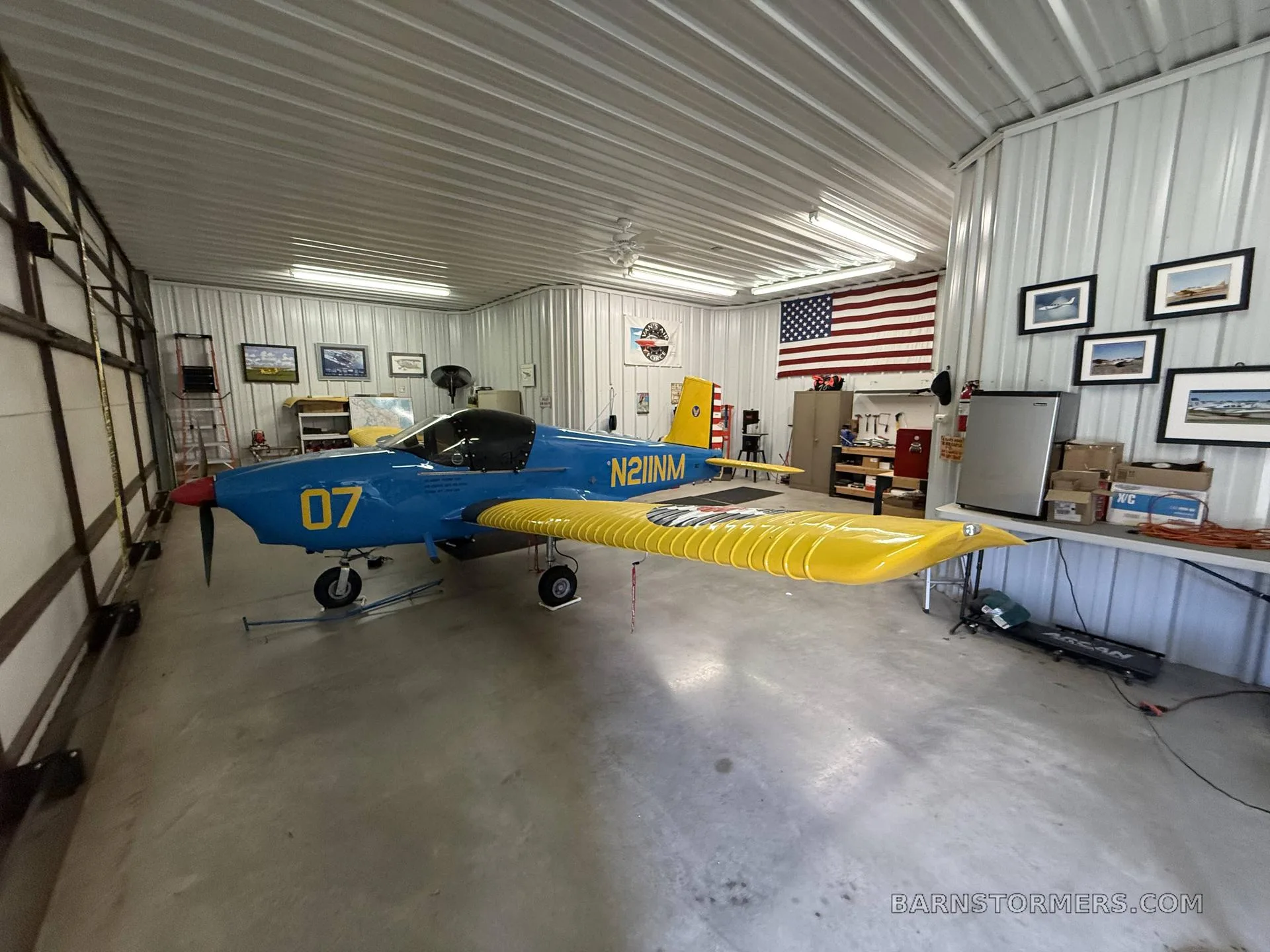 A blue and yellow single-engine aircraft with '07' on the nose and 'N211NM' on the fuselage, parked in a hangar.