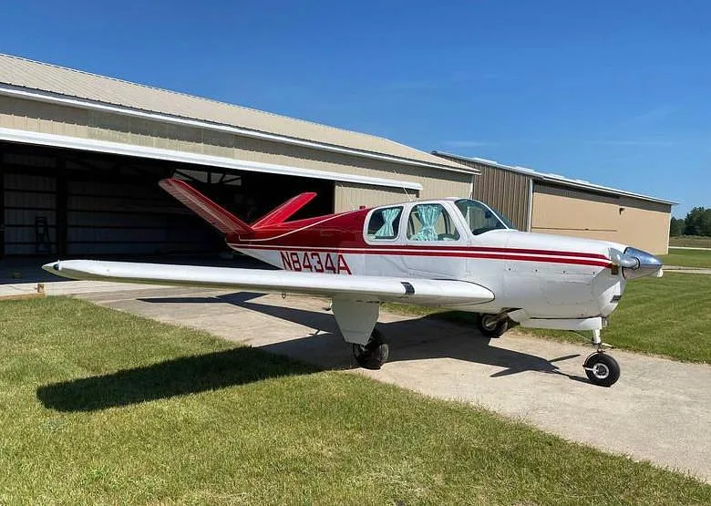 A white and red single-engine aircraft, likely a Beechcraft Bonanza, is parked on a tarmac in front of a hangar under a clear blue sky.