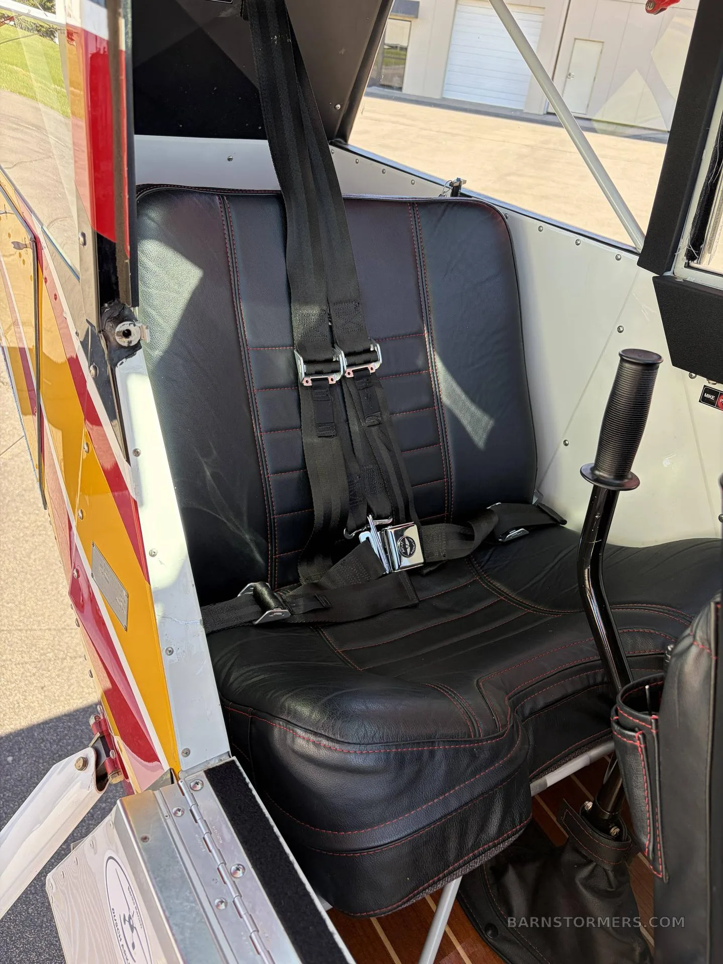A close-up of a black leather aircraft seat with red stitching and a five-point harness, seen from the side.