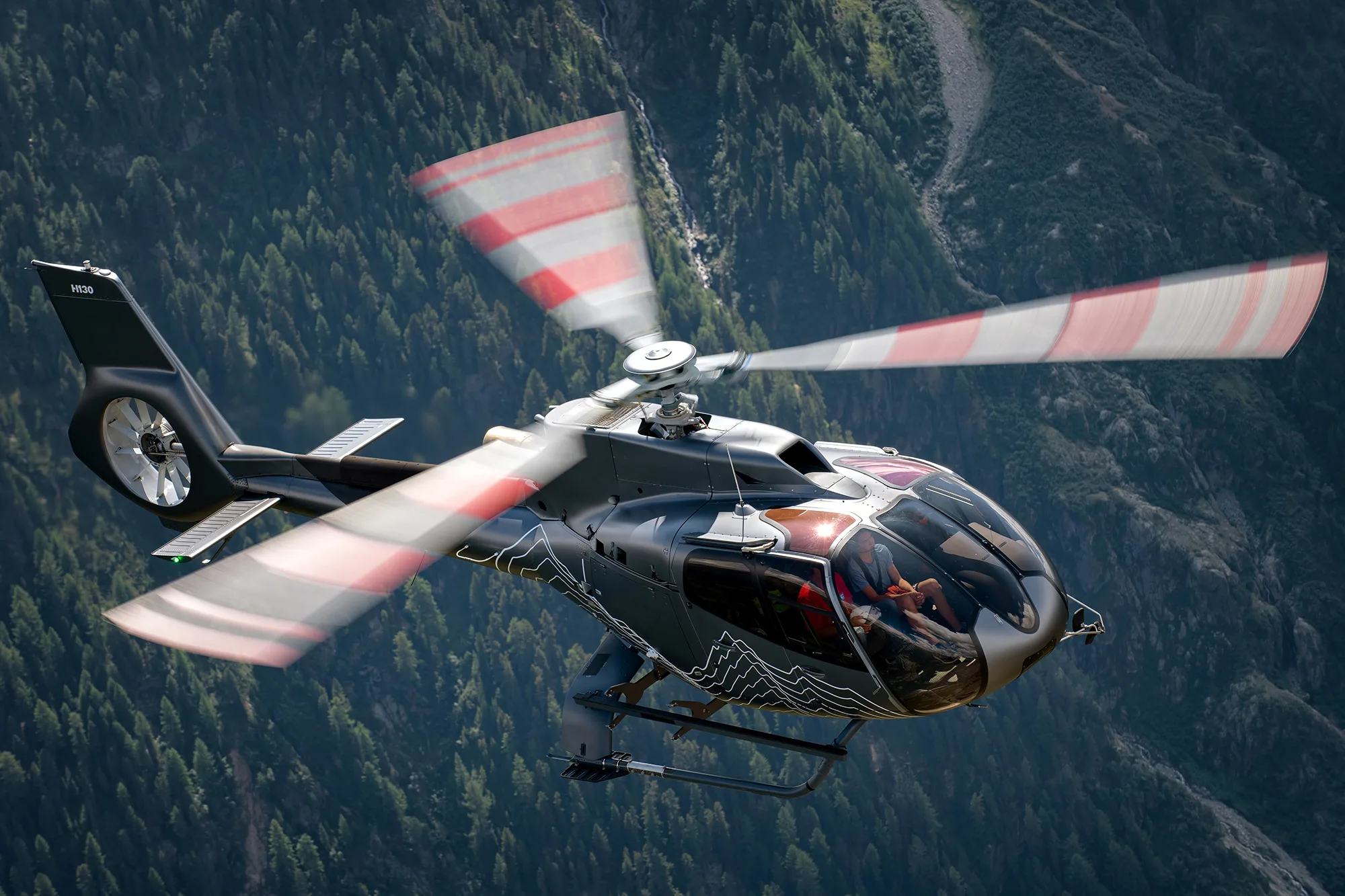 An Airbus H130 helicopter with a dark grey and white mountain-themed paint scheme flies over a forested mountain range.