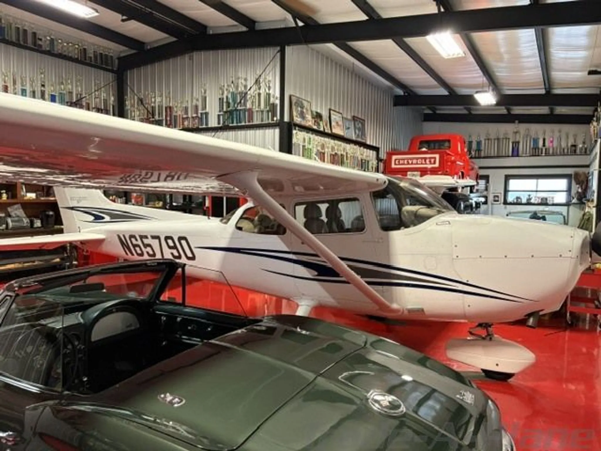 A white Cessna N65790 with blue and black stripes is parked in a hangar next to a green classic car and a red pickup truck.