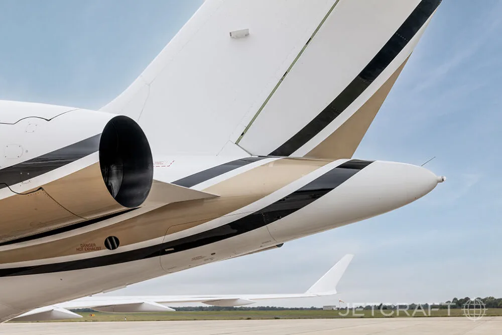 A close-up view of the tail section of a white Bombardier Global business jet, showing the engine and gold and black stripes.
