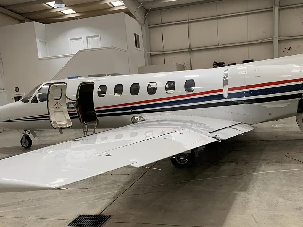 A close-up side view of a white business jet with red and blue stripes, showing the open main door and wing inside a hangar.