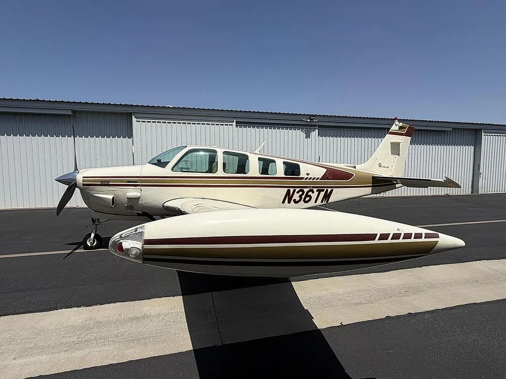 A white and gold Beechcraft Bonanza N36TM with tip tanks parked on an asphalt ramp next to hangars.