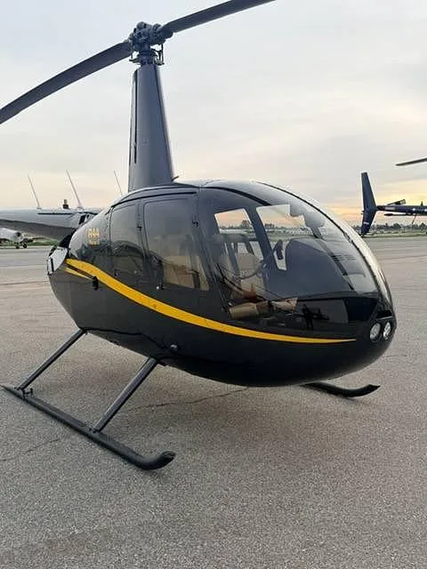 A black Robinson R44 helicopter with a yellow stripe, viewed from the front-right on an airfield.