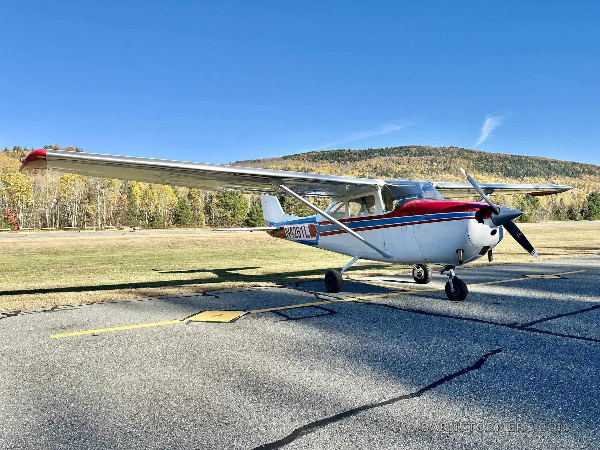 A white single-engine propeller aircraft with red and blue stripes, viewed from the front-right, parked on an asphalt ramp with autumn trees and mountains in the background.