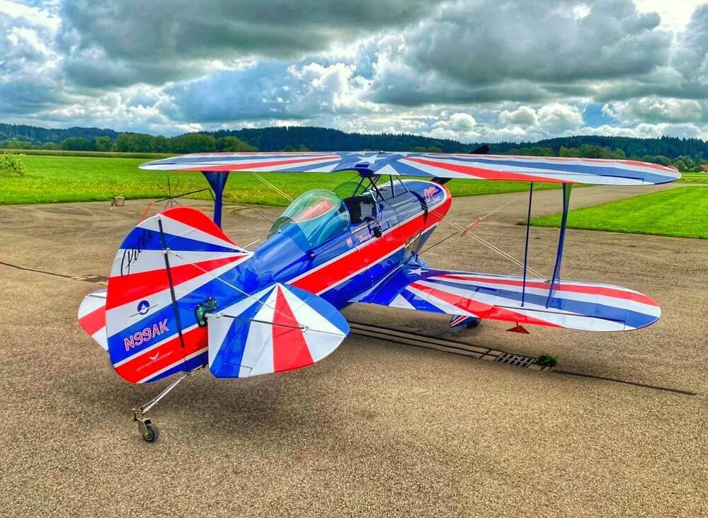 A red, white, and blue striped biplane, N99AK, sits on a tarmac under a cloudy sky.
