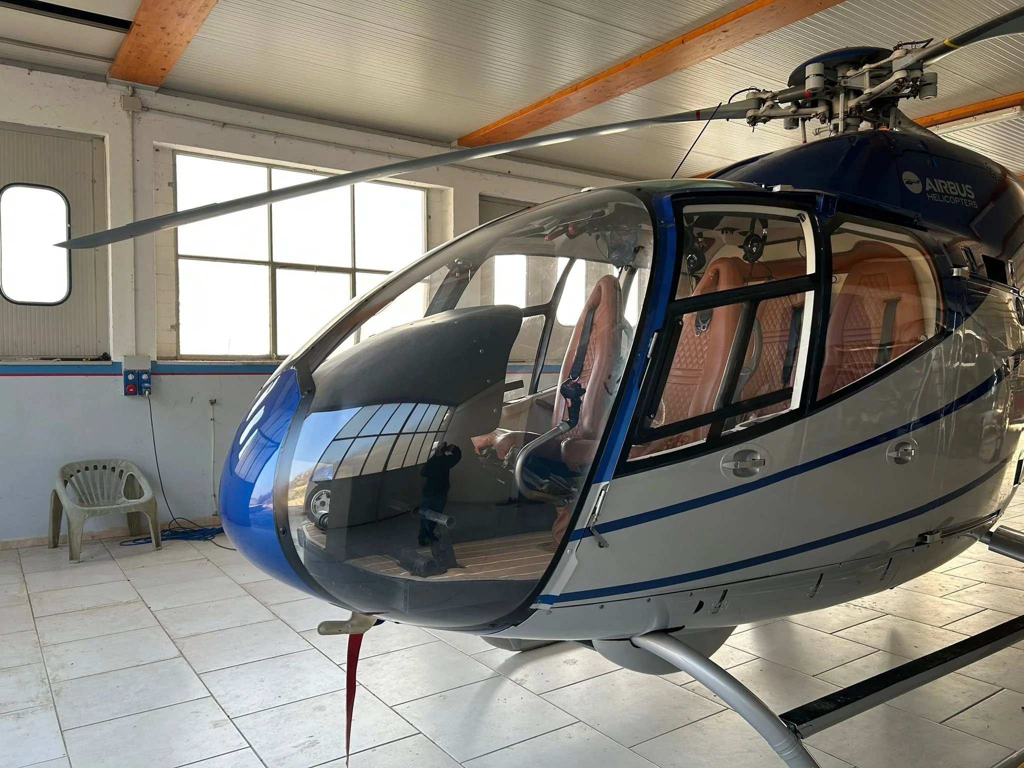 Front-side view of an Airbus H120 Colibri helicopter with blue and silver livery inside a hangar, showing the cockpit and brown leather seats.
