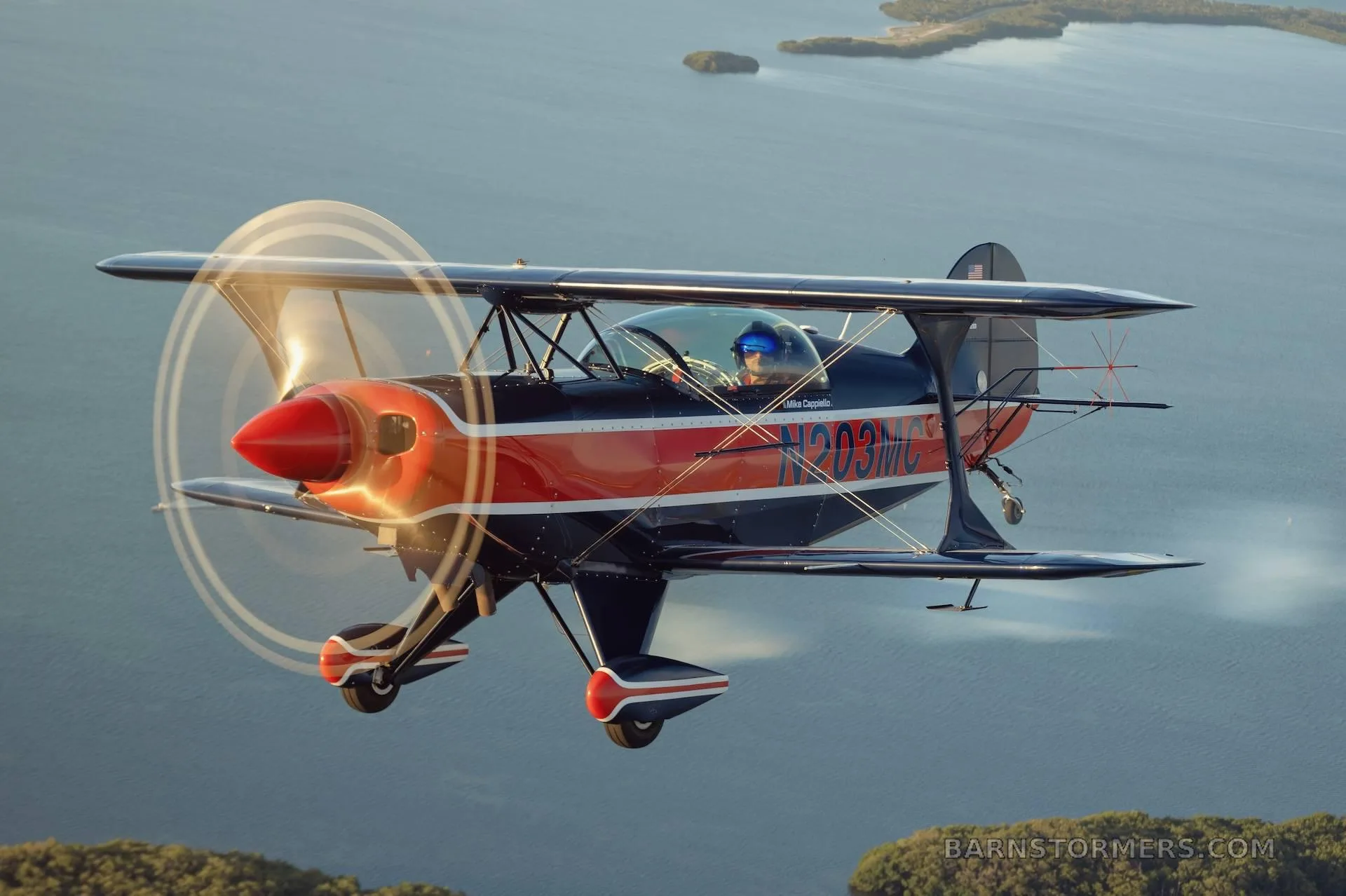 A dark blue and red biplane with registration N203MC flies over a body of water with islands.