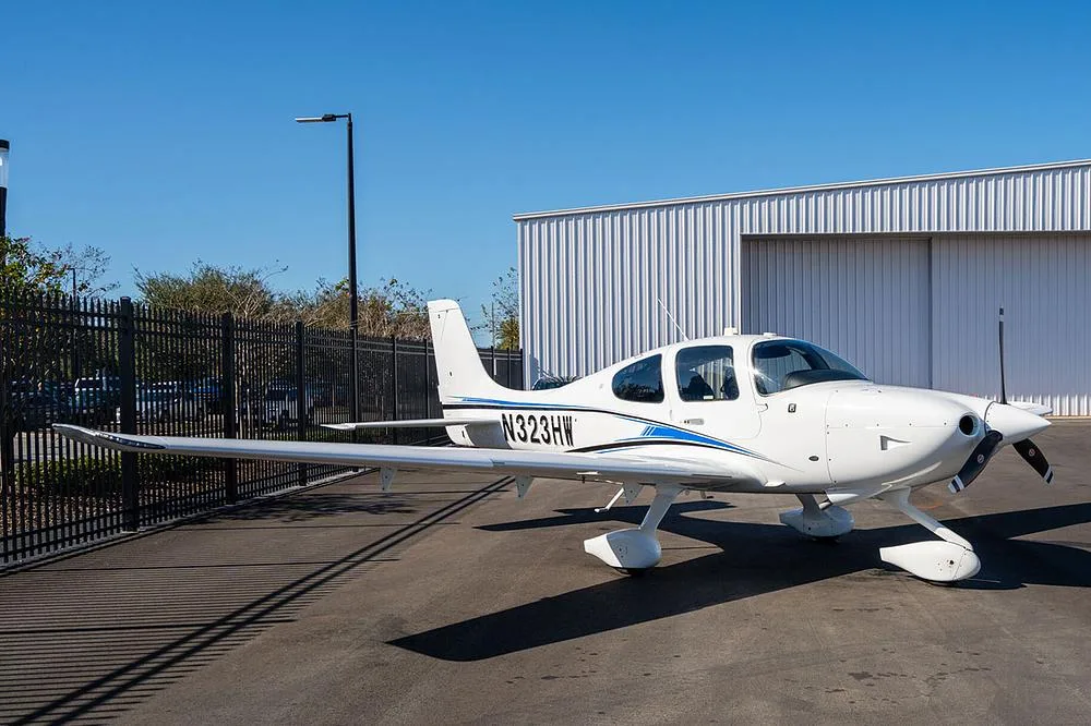 A white Cirrus SR22 aircraft with blue and black stripes, registration N323HW, parked on an asphalt ramp next to a white metal hangar.