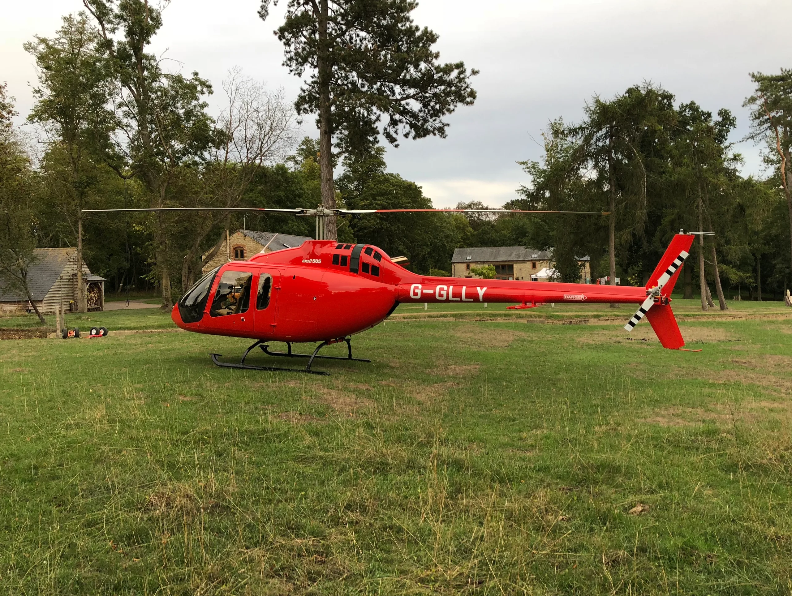 A red Bell 505 helicopter, registration G-GLLY, sits on a grassy field with buildings in the background.