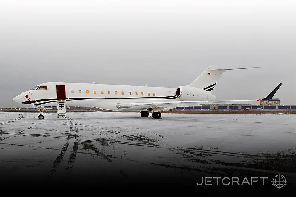 A white Bombardier Global Express business jet is parked on a snowy tarmac with its entry door open.