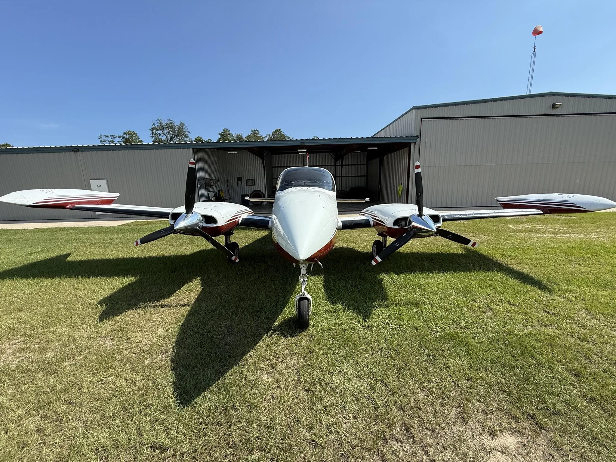A white and red twin-engine propeller aircraft parked on a grassy field in front of a hangar on a sunny day.