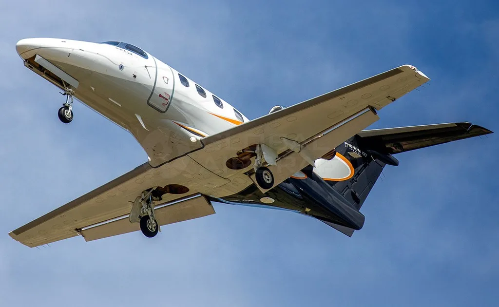 An Embraer Phenom 100 business jet with black and orange livery is seen from below, in flight with landing gear extended against a blue sky.