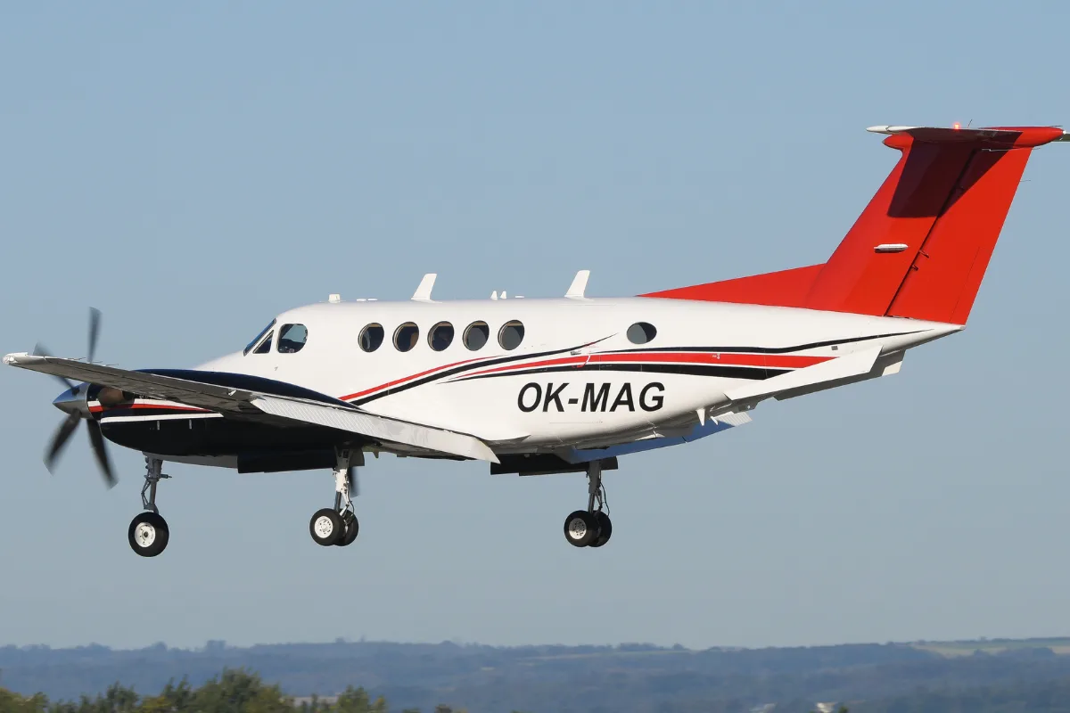 A white and red twin-engine turboprop aircraft with registration OK-MAG, landing against a clear blue sky.