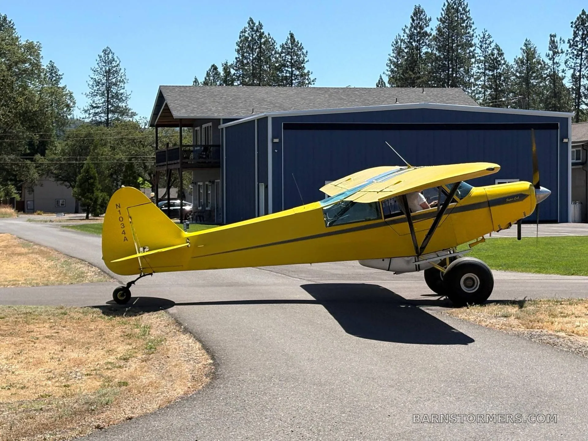 A bright yellow Piper Super Cub aircraft with large tundra tires is parked on an asphalt path in front of a blue hangar on a sunny day.