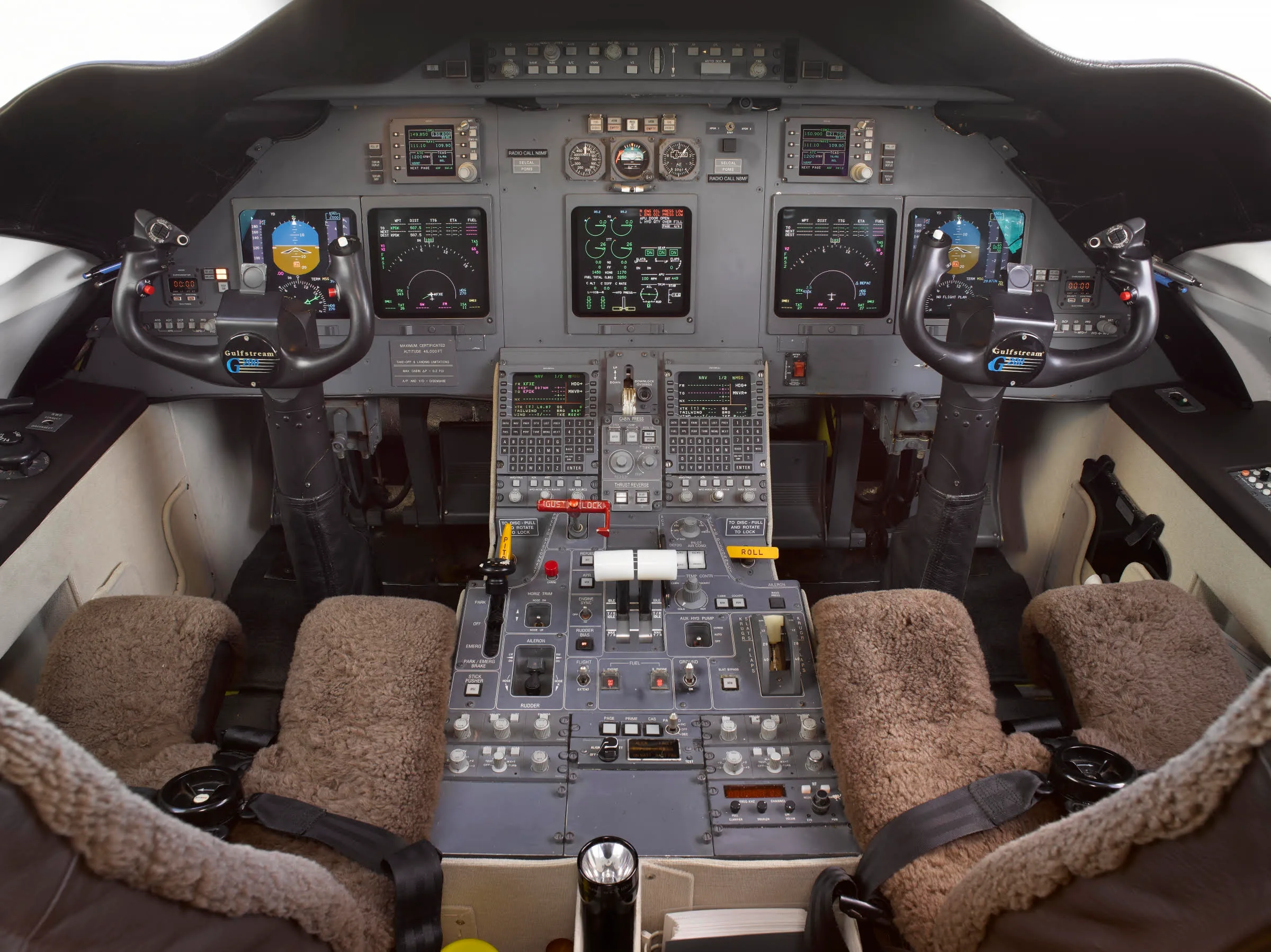 The cockpit of a Gulfstream aircraft displays multiple screens, control yokes, and a central throttle quadrant, with sheepskin seat covers.