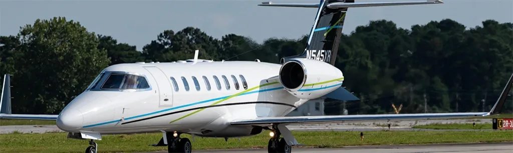 A white business jet with blue and green stripes and registration N545XR on its tail, taxiing on a tarmac with trees in the background.