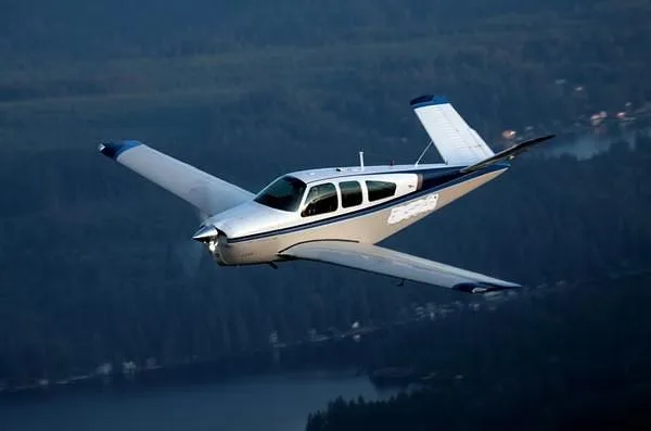 A silver and blue single-engine aircraft with a V-tail flies over a dark landscape with water and trees.