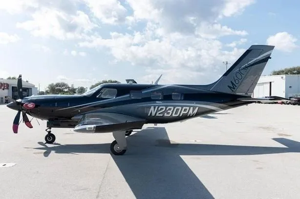 A side view of the black Piper M600 aircraft, registration N230PM, parked on a tarmac with buildings in the background.