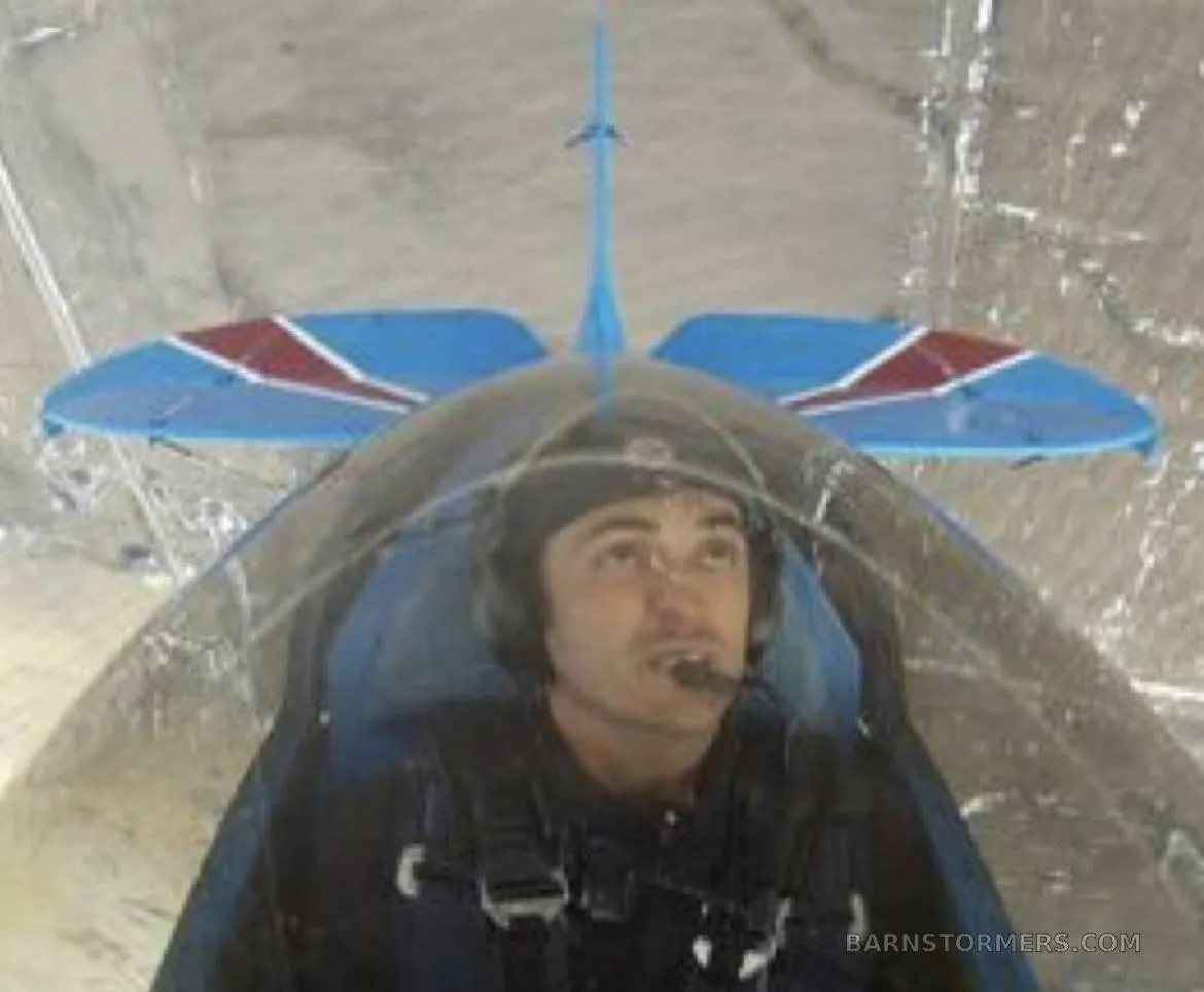 A pilot in a helmet and headset looks up from the cockpit of a blue and red biplane.