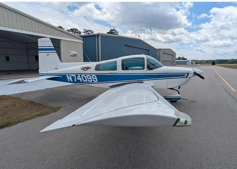 A side view of the white and blue Grumman AA-5 Traveler N74099 parked on an airfield with hangars in the background.