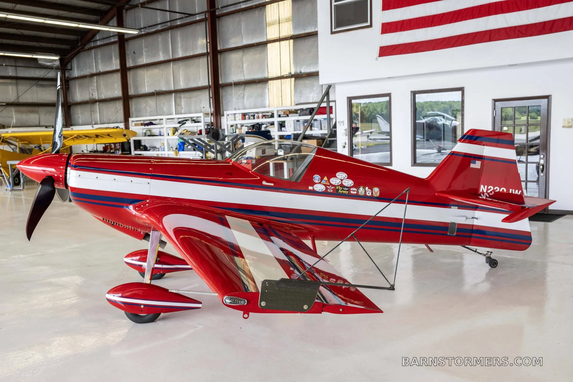 A red and white single-engine aerobatic aircraft with blue stripes, seen from the side in a hangar.
