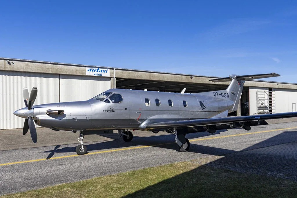 A silver Pilatus PC-12 aircraft with registration OY-GSA parked on an airfield next to a hangar.
