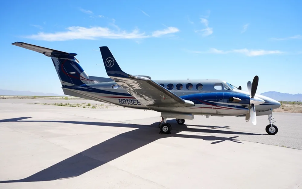 A silver and blue twin-engine turboprop aircraft (likely a King Air) parked on a tarmac with mountains in the background under a blue sky.