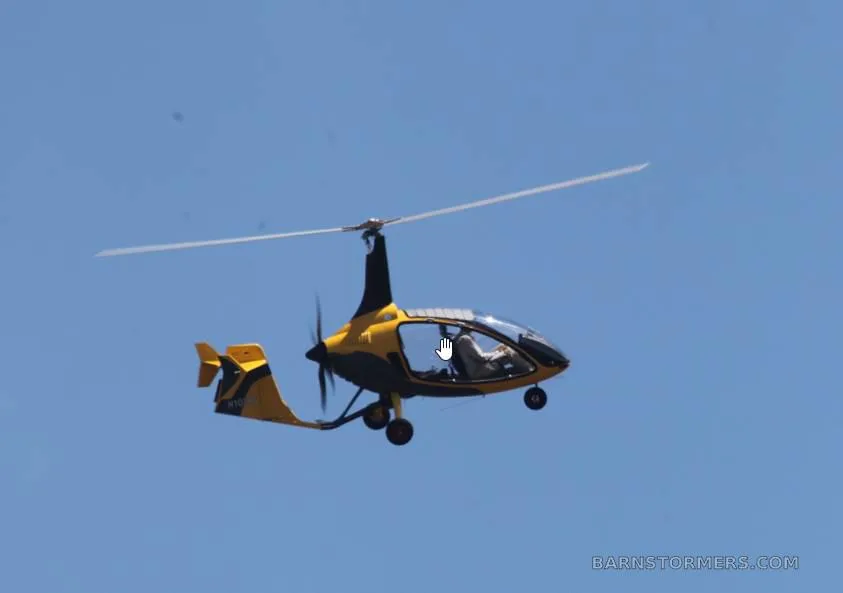 A yellow and black gyrocopter with a pilot inside, flying against a clear blue sky.