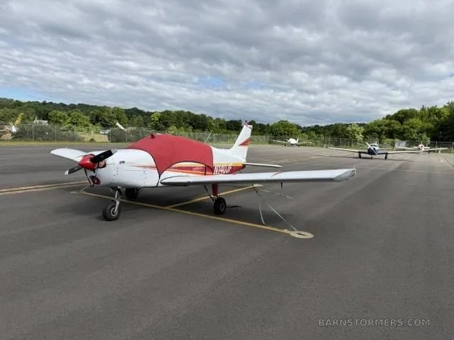 A white and red single-engine aircraft with a red engine cover parked on an asphalt apron under a cloudy sky.