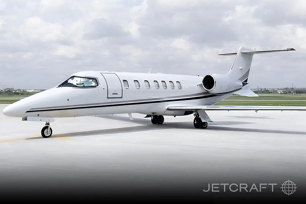 A white business jet with black and grey stripes is parked on a tarmac under a cloudy sky.