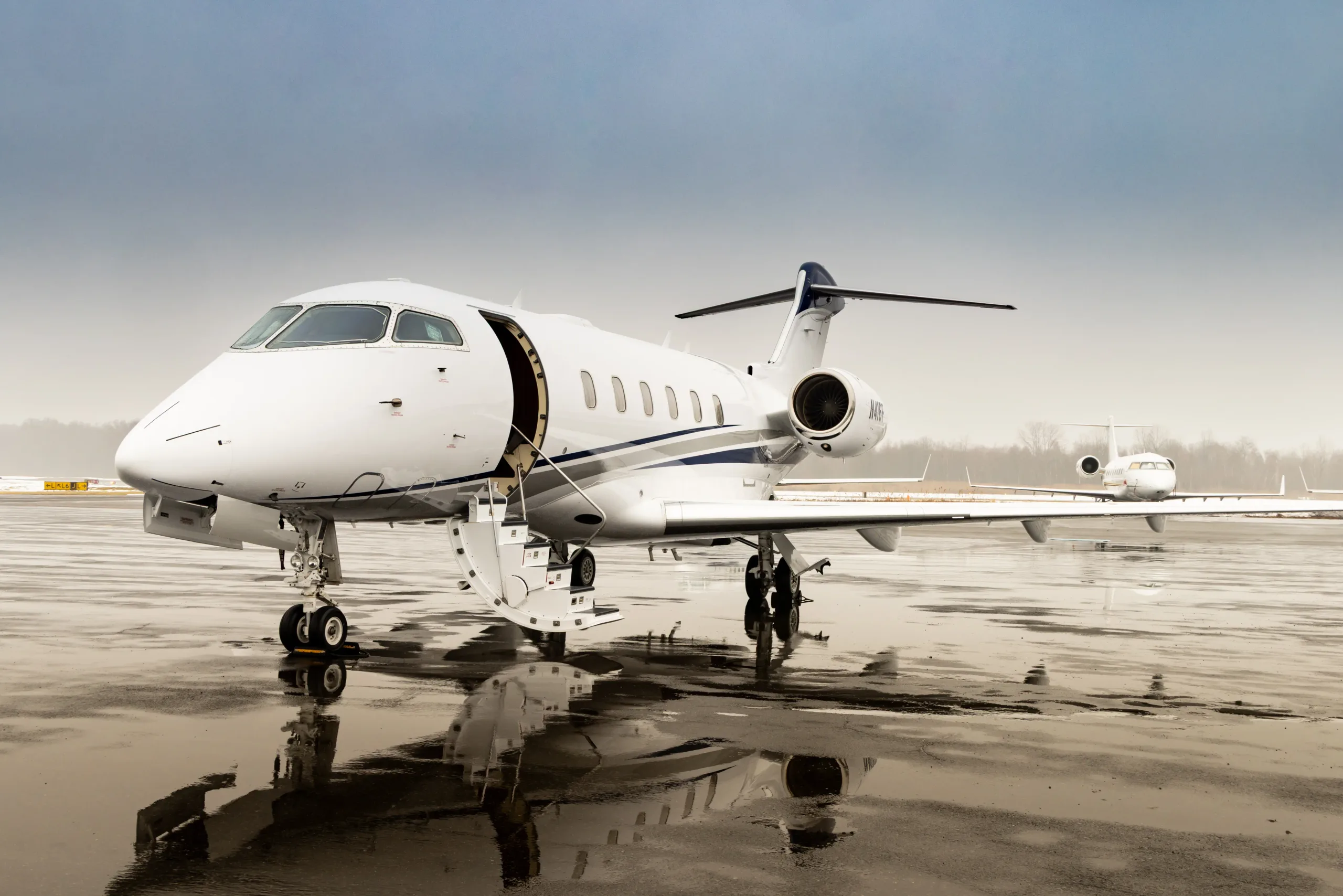 A white Bombardier Challenger 300 business jet with blue and grey stripes on a wet tarmac, with its main door open.