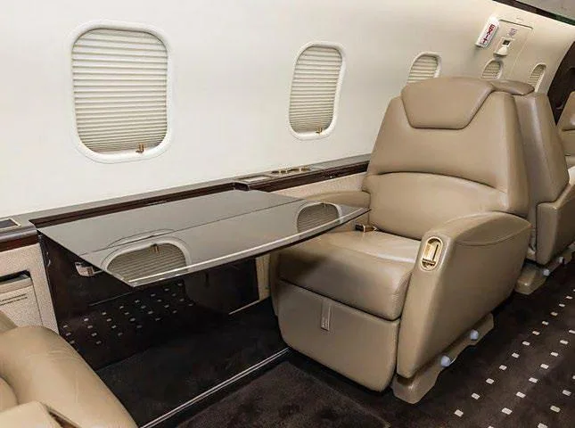 A close-up of a beige leather cabin seat with a polished wooden side table extended, next to a window with blinds.
