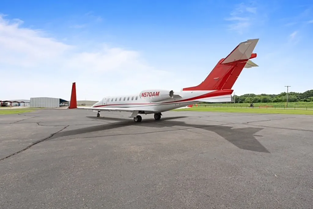 A white and red business jet (N570AM) parked on an asphalt tarmac with an open field in the background.