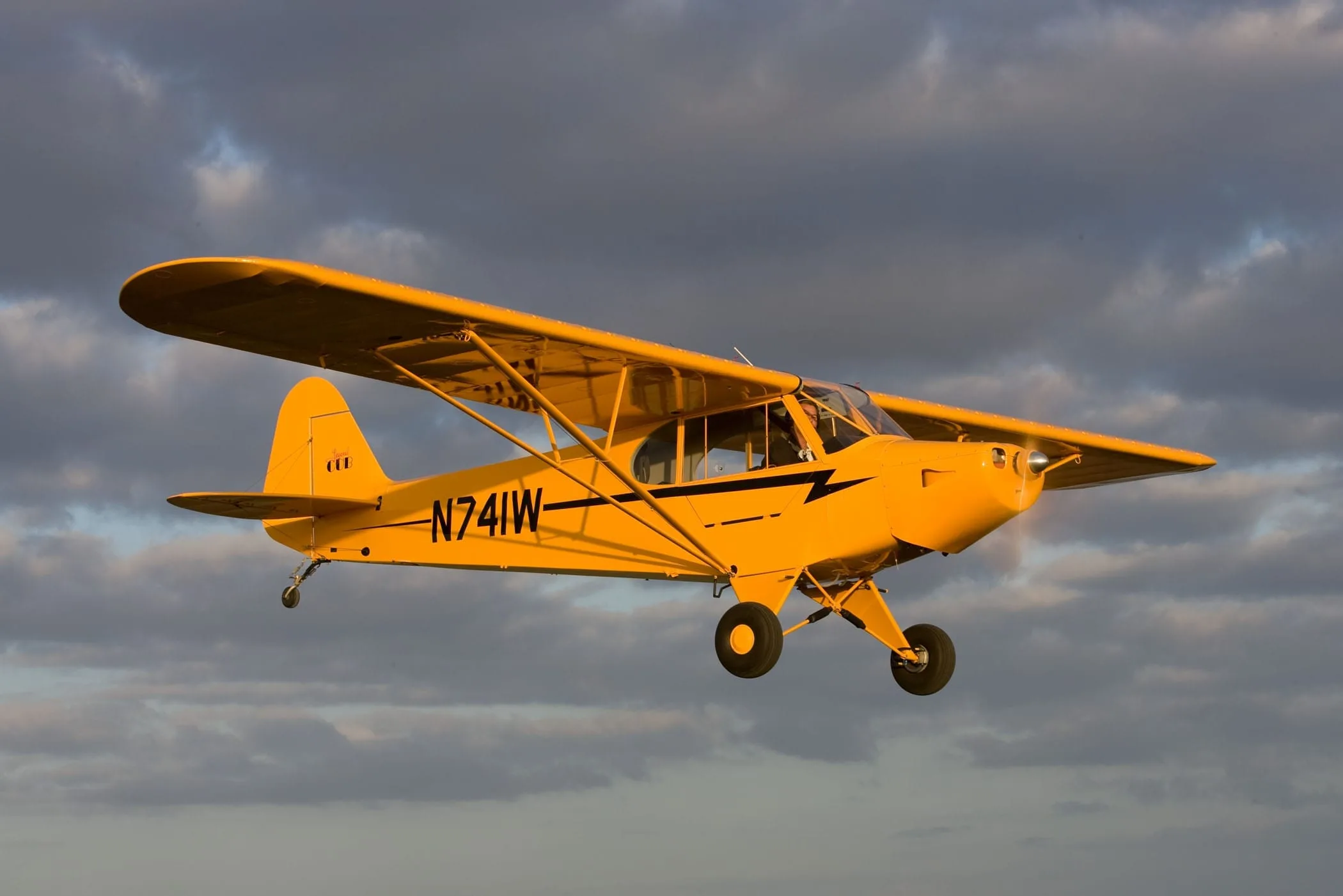 A yellow Piper Cub-style aircraft (N741W) with black stripes flies against a dramatic cloudy sky.