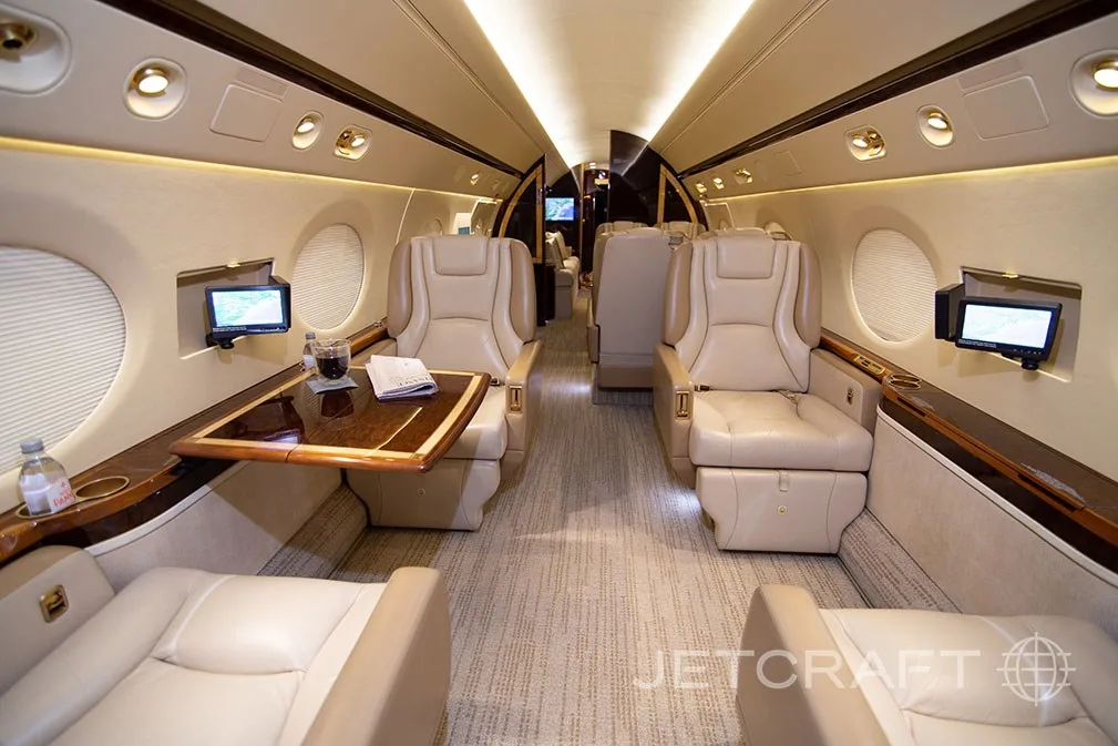 A private jet cabin with beige seats, a wooden table, and individual entertainment screens.