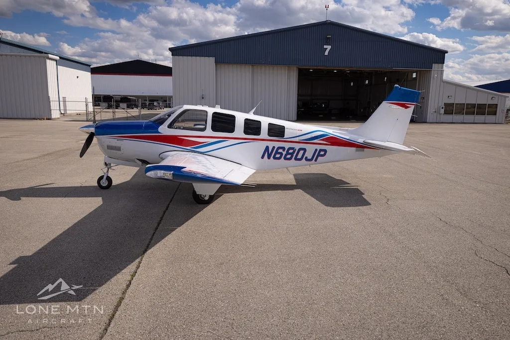 A side view of a white, blue, and red Beechcraft Bonanza N680JP parked on an airfield next to a hangar.