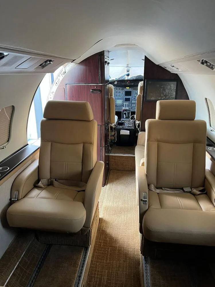 View from the aircraft cabin showing two tan leather seats and dark wood paneling, looking forward towards the cockpit.