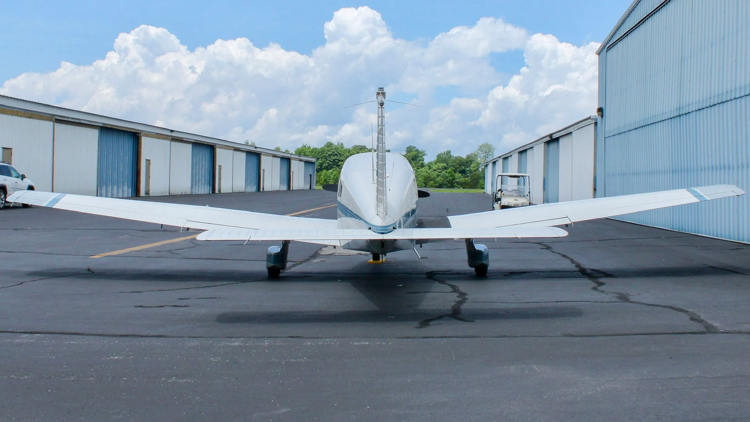 A rear view of a white and blue Piper Cherokee aircraft parked on an asphalt ramp between two rows of hangars.