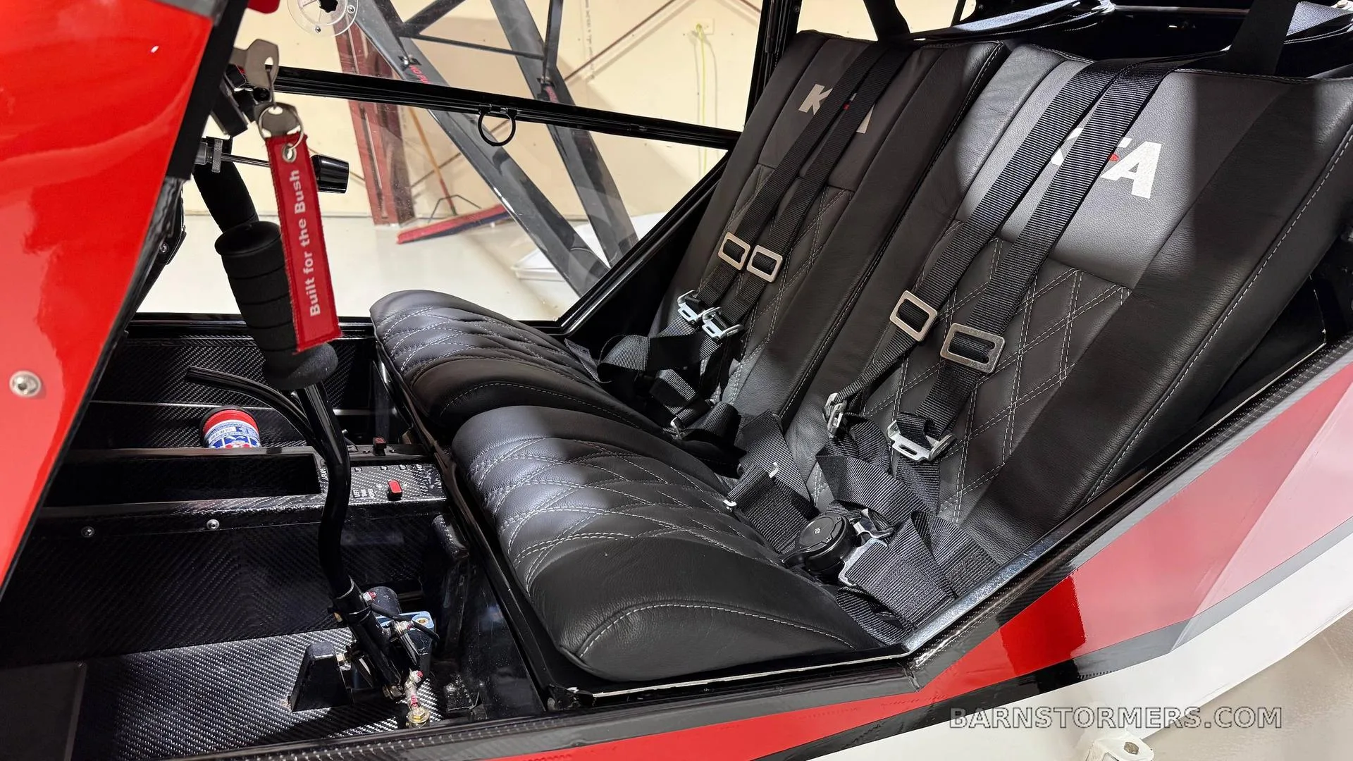 The black leather two-seat interior of a Kitfox aircraft, showing harnesses and a "Built for the Bush" keychain.
