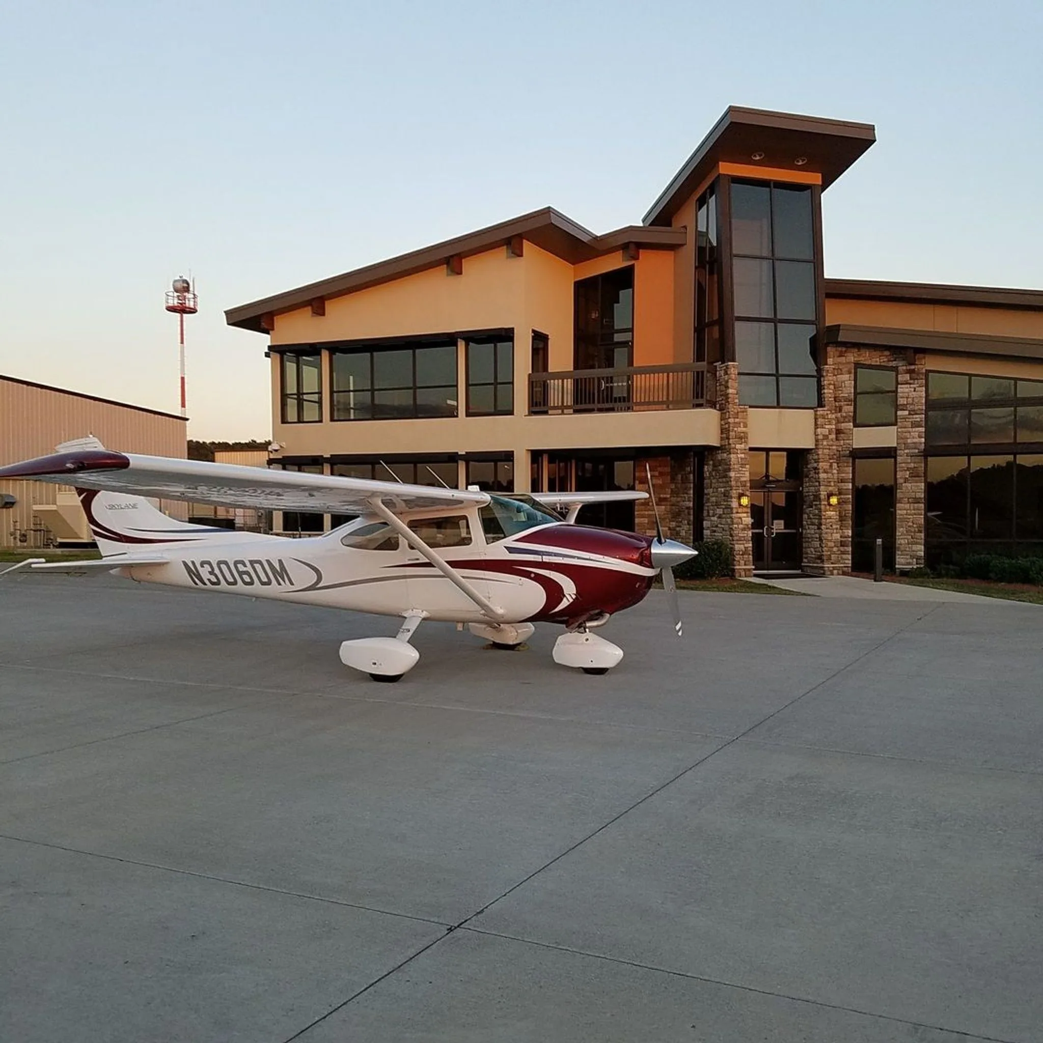 A white and maroon Cessna aircraft, N306DM, parked on an apron in front of a modern airport building at sunset.