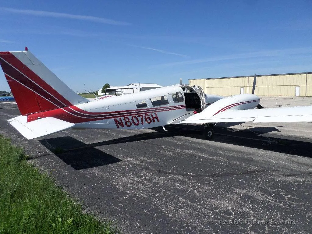 A white and red single-engine propeller aircraft N8076H with its door open, parked on an asphalt ramp next to hangars.