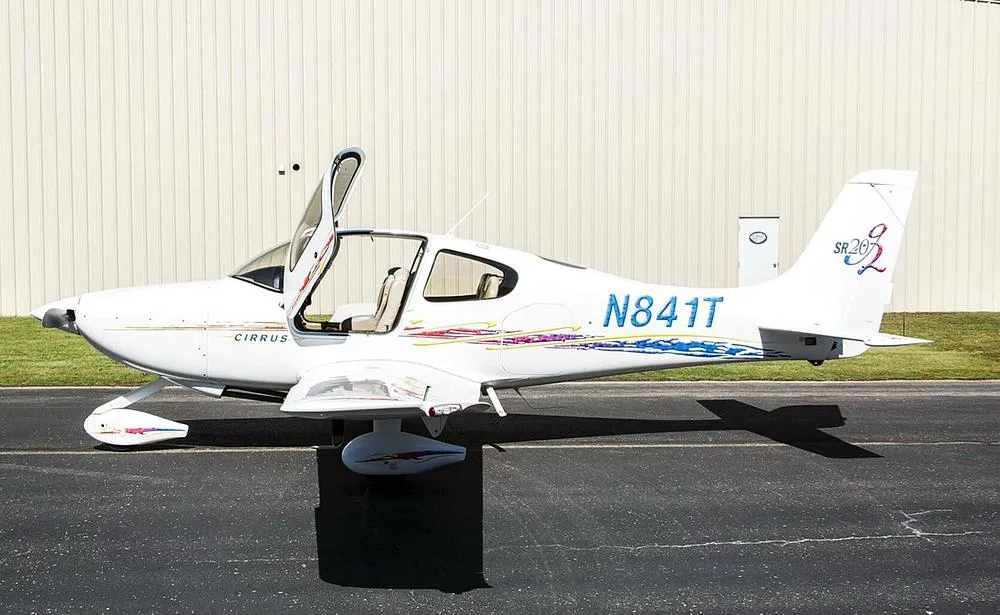 A white Cirrus SR20 aircraft with open doors and blue and red accent stripes parked on an airfield.