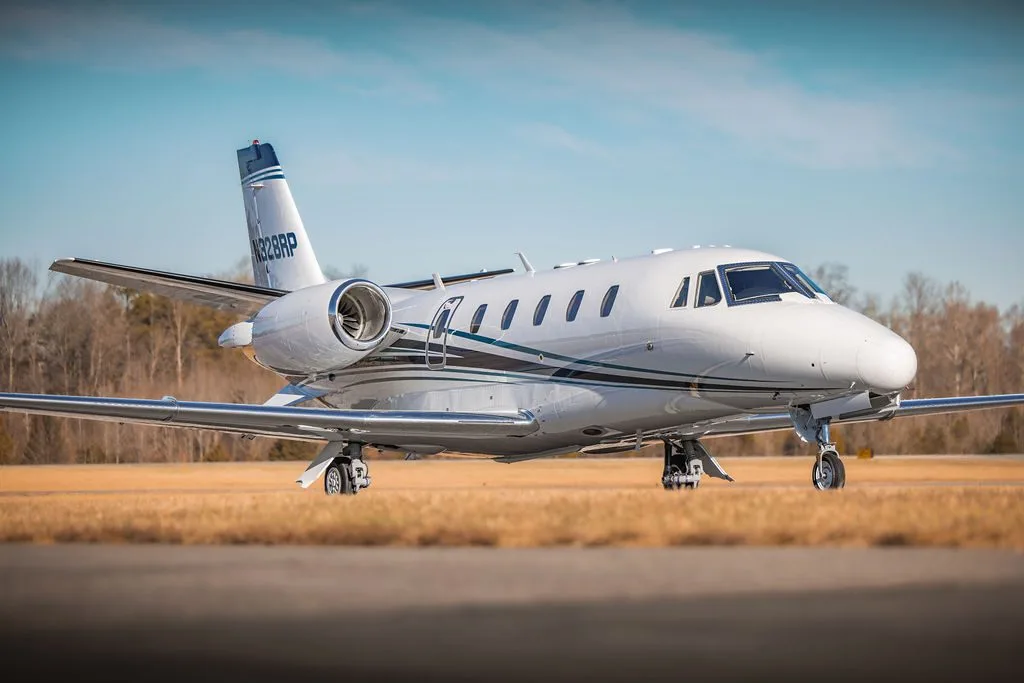 A white business jet with blue and black stripes, registration N328RP, parked on an airfield with dry grass and trees in the background.