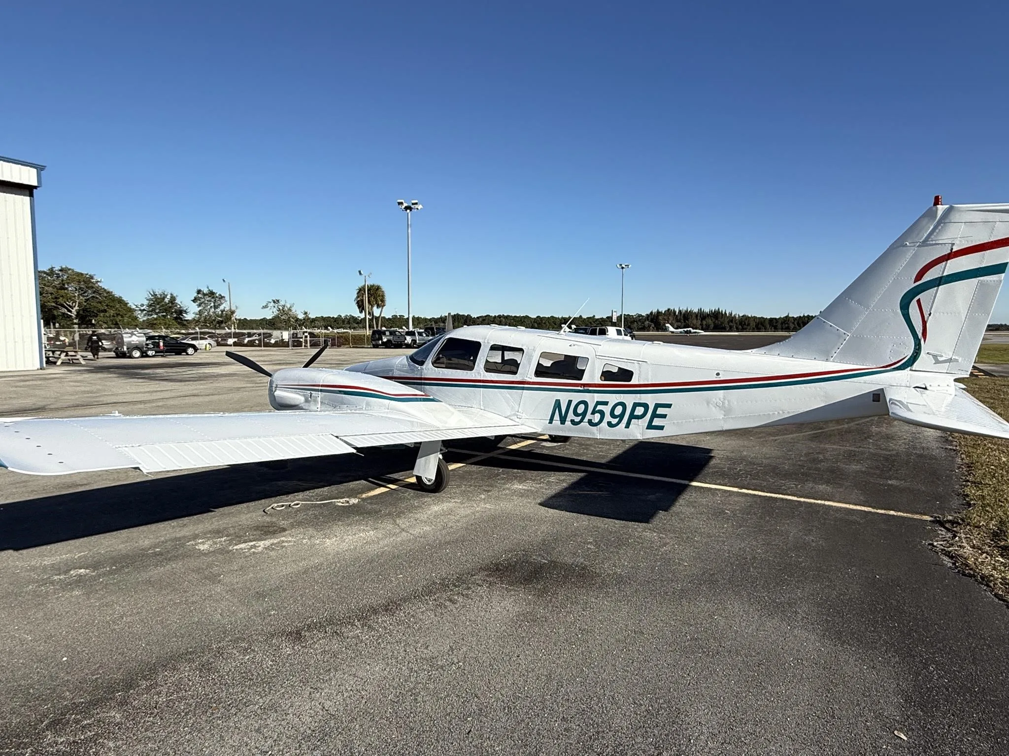 A white twin-engine Piper Seneca II, N959PE, with teal and red stripes, parked on an airport tarmac next to a hangar.