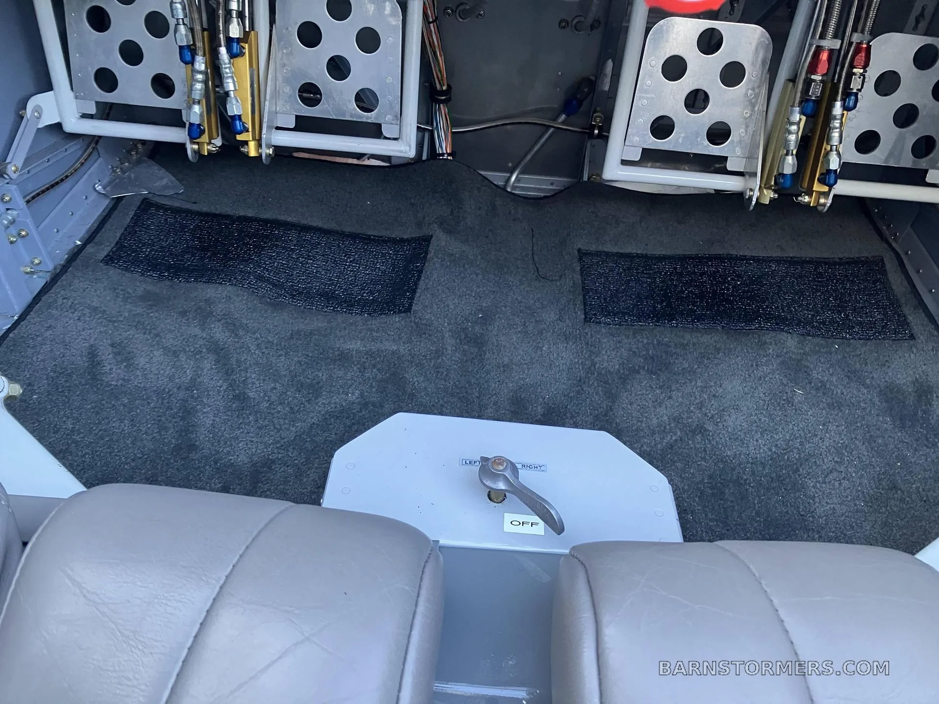 A view of the aircraft's grey carpeted floor, rudder pedals, and a fuel selector switch between two light grey seats.