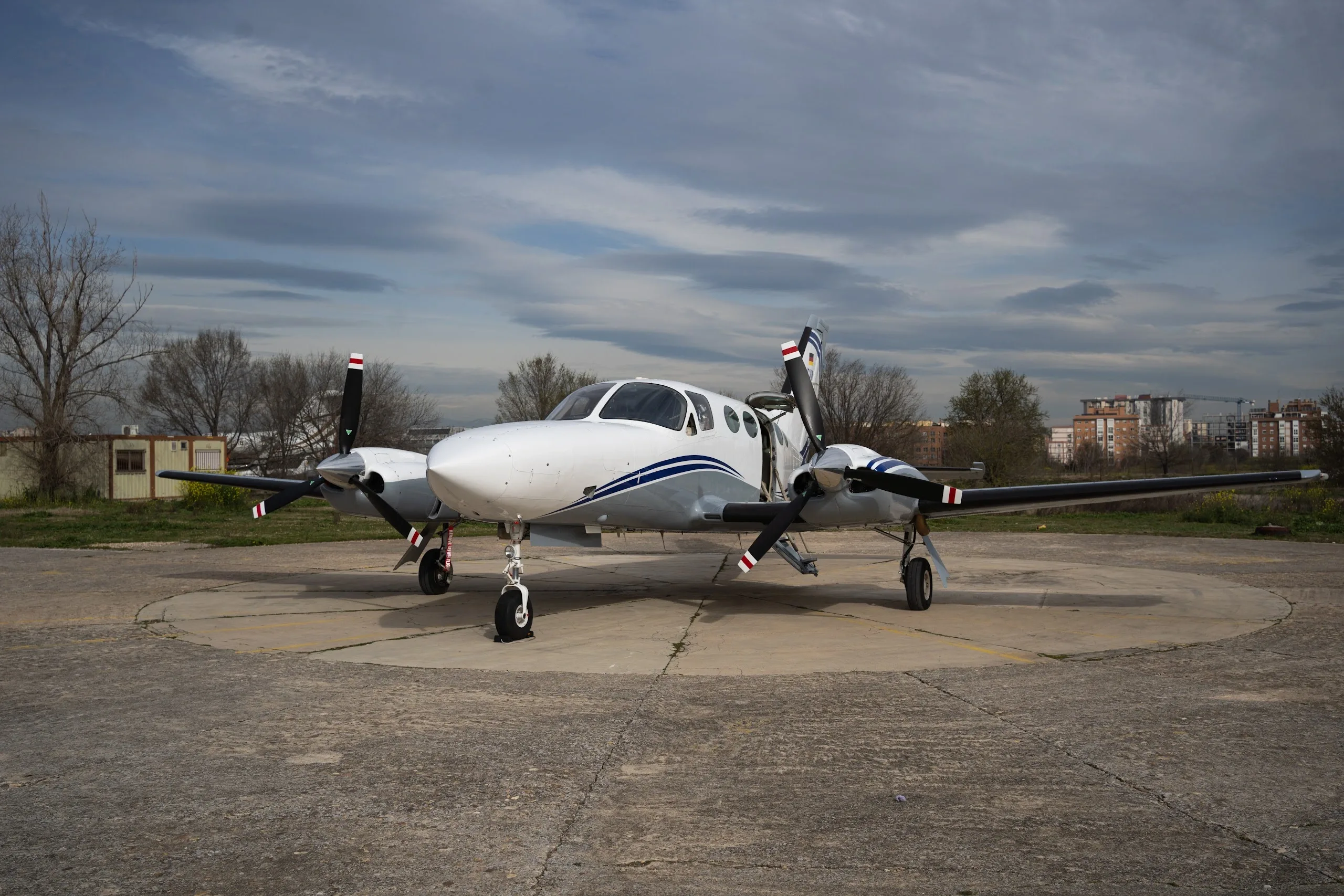 A white and grey twin-engine propeller aircraft with blue stripes on a concrete pad under a cloudy sky.